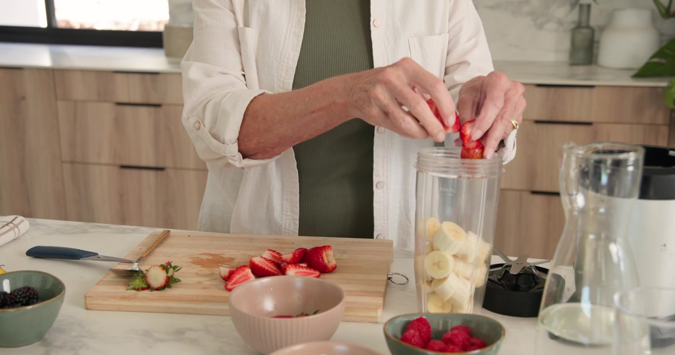 Donna anziana che seleziona ingredienti da una ciotola, affetta con un coltellino in cucina e riempie il frullatore per un frullato.