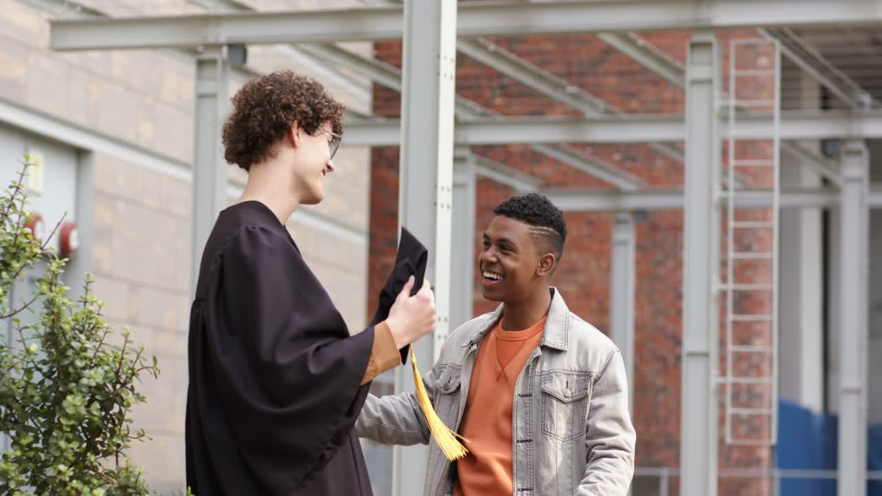 Diverse male friends shaking hands exchanging cap and examining tassel for graduation on campus