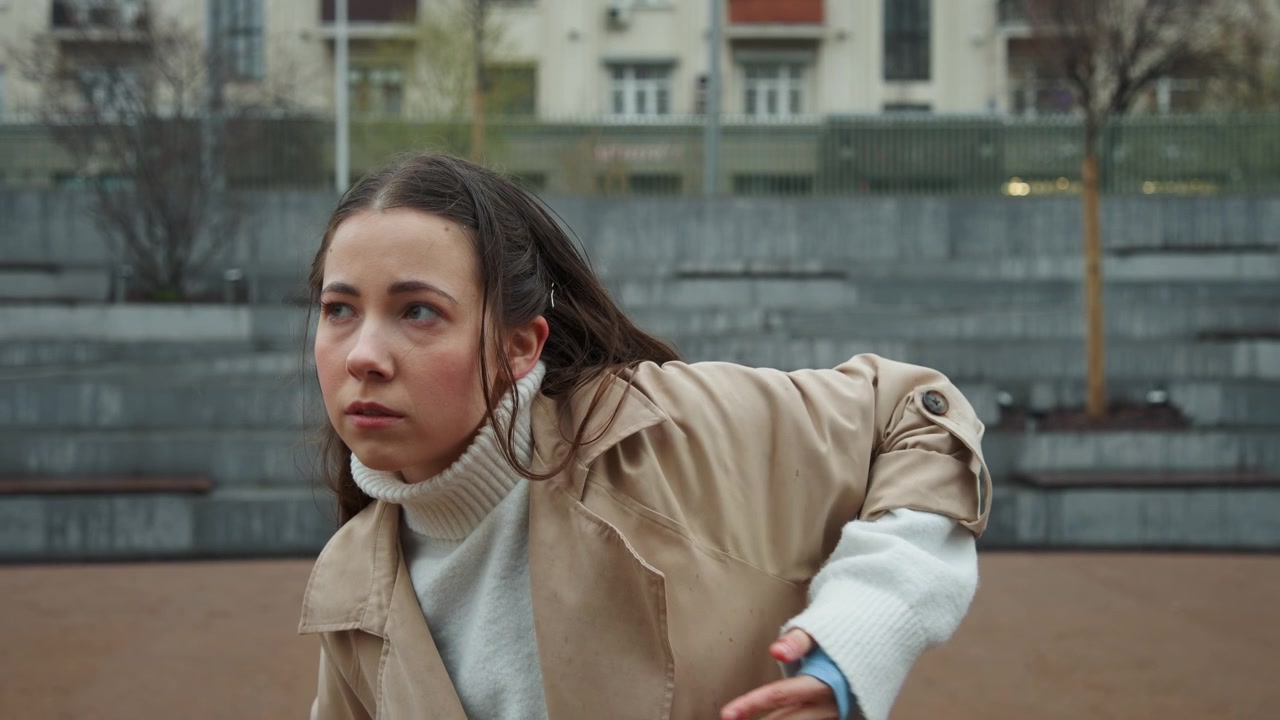 Primo piano di giovane donna che esegue movimenti di arti marziali all'aperto davanti a scalini di cemento e edifici. La donna indossa un trench beige sopra un dolcevita bianco con segni rossi visibili su di lei.