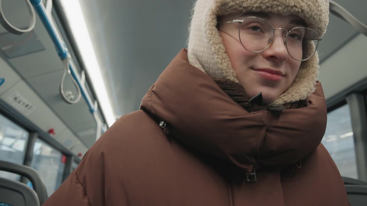 Young woman in thick winter coat with fur-trimmed hood and knit hat stands inside near-empty tram, holding blue overhead rail with steady grip, relaxed expression, and quiet urban commute mood