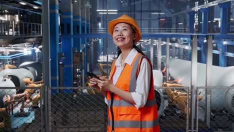 vista lateral de una ingeniera asiática con casco de seguridad usando un teléfono inteligente y mirando a su alrededor mientras está de pie en una fábrica de fabricación de turbinas eólicas