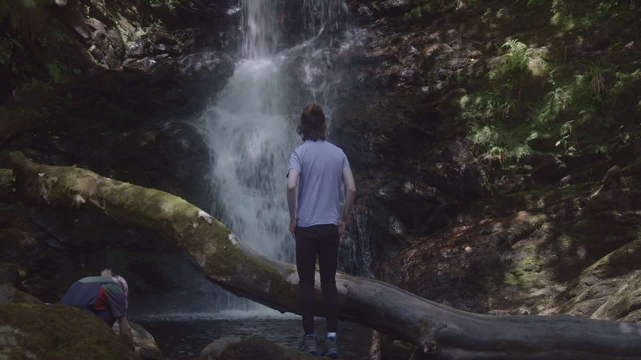 전경에 큰 나무 줄기가 있는 스코틀랜드의 아름다운 폭포 앞에 있는 두 사람의 전망