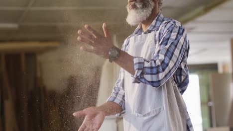 African-american-male-carpenter-wearing-an-apron-dusting-his-hands-at-a-carpentry-shop