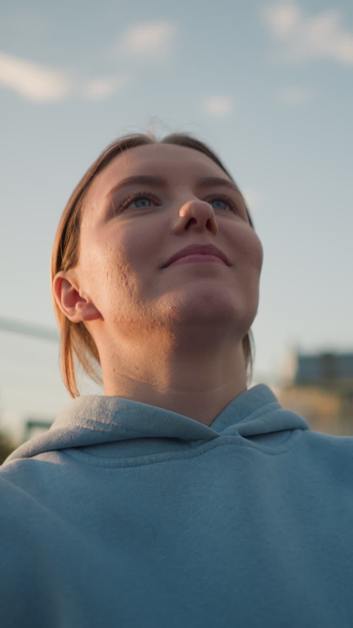 Close-up of lady in blue sweater playing volleyball outdoors, arms raised with a joyful expression, greenery and buildings create a scenic background under a bright sky