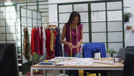 happy african american female fashion designer wearing tape measure looking at plans at office