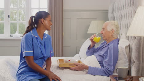 female care worker in uniform bringing senior man at home breakfast in bed on tray