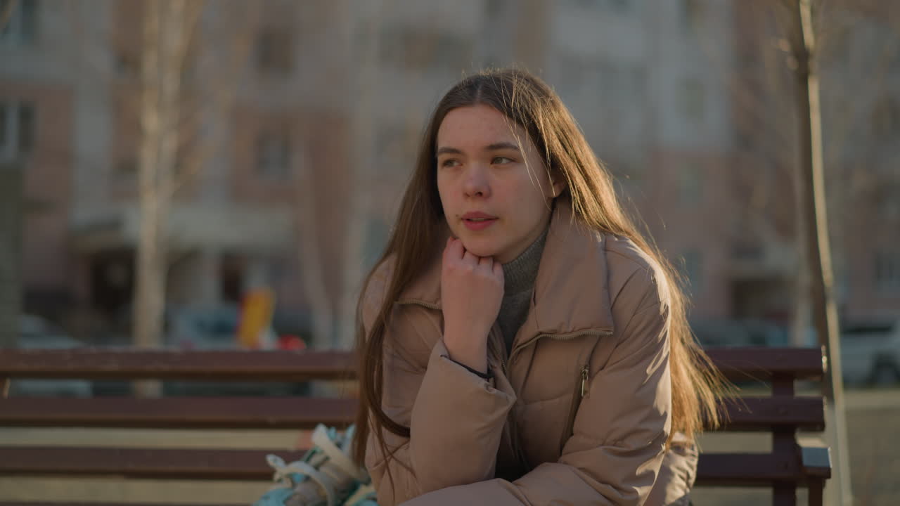 A young girl in a peach jacket sits in a park, deep in thought with her chin resting on her hand. Her expression is reflective, as she meditates quietly in the peaceful outdoor setting