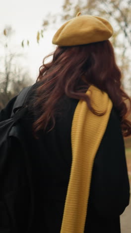vista trasera de una mujer con un silenciador amarillo y una boina caminando por el bosque, sosteniendo el silenciador mientras mira hacia un lado, con el cabello balanceándose suavemente en la brisa de otoño