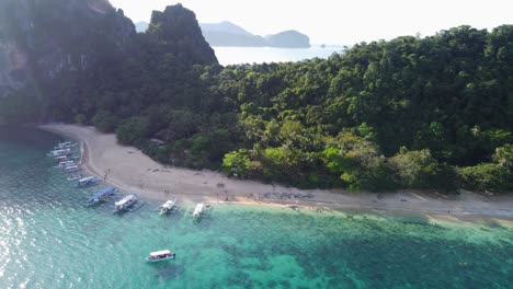 Aerial-view-:-multiple-filipino-banca-tour-boats-anchored-in-turquoise-clear-water-along-white-sand-beach-against-tropical-beach-hut-of-Helicopter-Island,-El-Nido,-People-walking-and-swimming