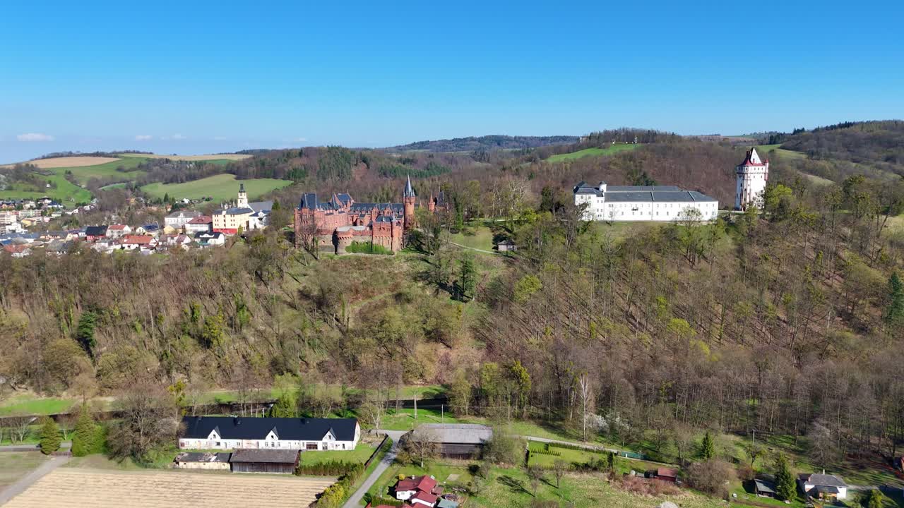 Drone em voo reverso revela o castelo de Hradec nad Moravicí no topo de uma colina, com a paisagem campestre, a vila e o relevo ondulado ao redor, sob um céu azul límpido.
