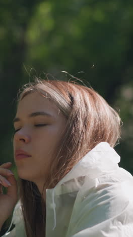 joven se quita la capucha del impermeable, descansando la mano en la barbilla, perdida en pensamientos, la suave luz del sol ilumina su expresión pensativa contra un fondo verde suavemente borroso