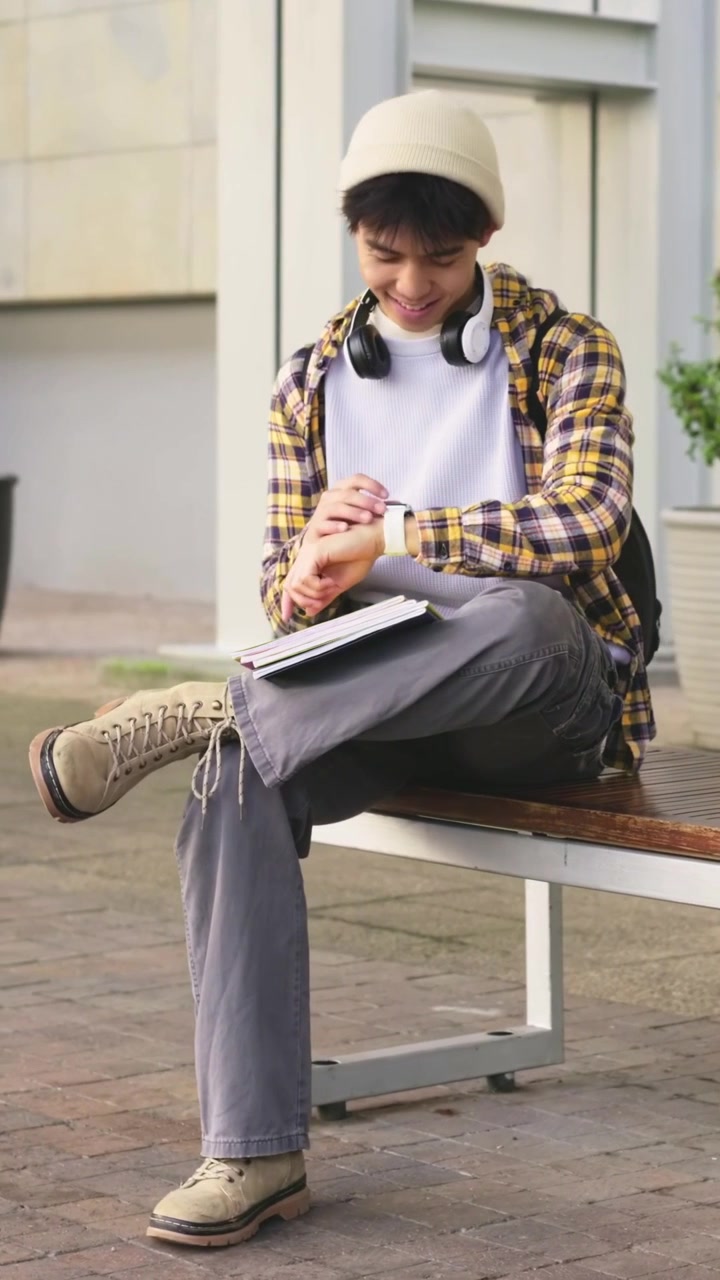 Video verticale: Studente con berretto che guarda di sfuggita e tocca lo smartwatch su una panchina in piazza, con un quaderno