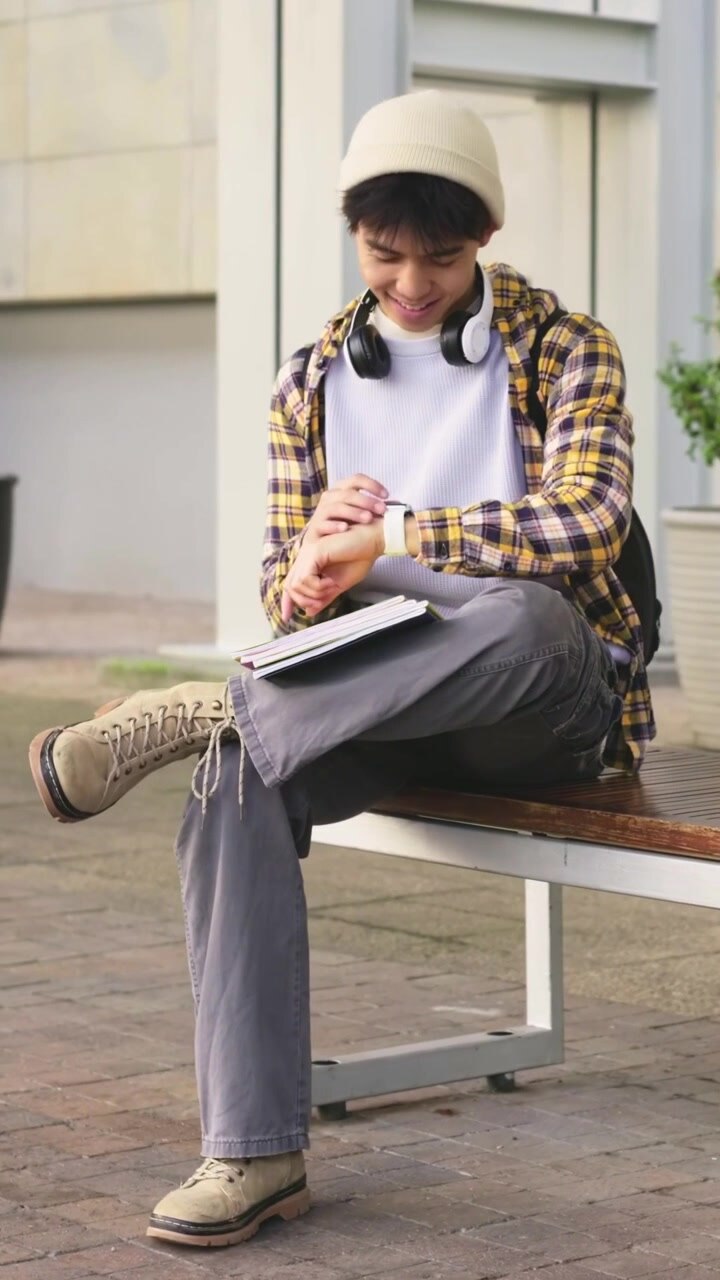 Video verticale: Studente con berretto che guarda di sfuggita e tocca lo smartwatch su una panchina in piazza, con un quaderno