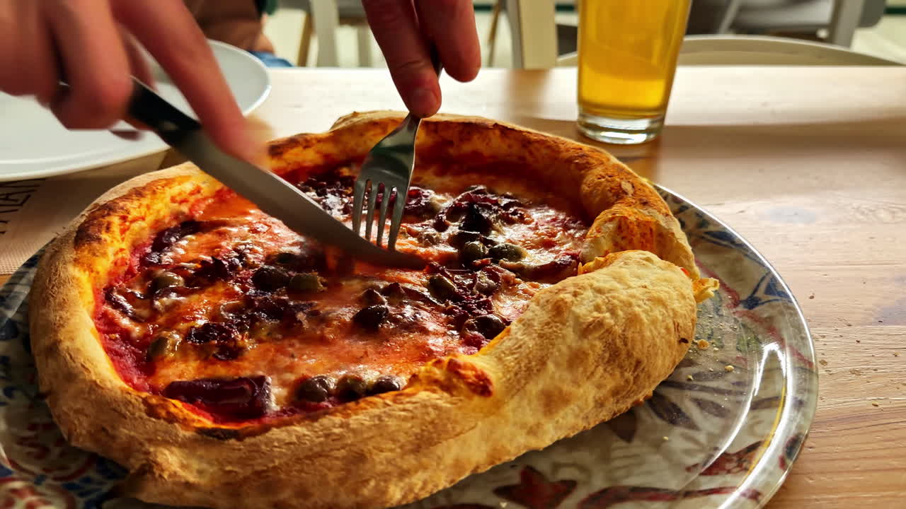 Riprese ravvicinate che catturano il momento in cui una pizza viene tagliata con coltello e forchetta a un tavolo di ristorante. L'azione del taglio rivela formaggio fuso, salsa di pomodoro e una crosta spessa in movimento