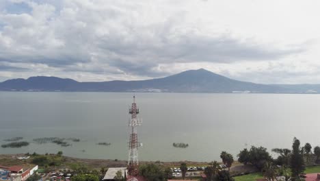 Travel-in,-moving-forward-drone-aerial-shot-of-a-huge-lake-near-a-small-town-with-an-antenna-in-front,-with-mountains-and-clouds-with-birds-crossing-flying-in-front-of-drone,-slow-motion,-slowmo