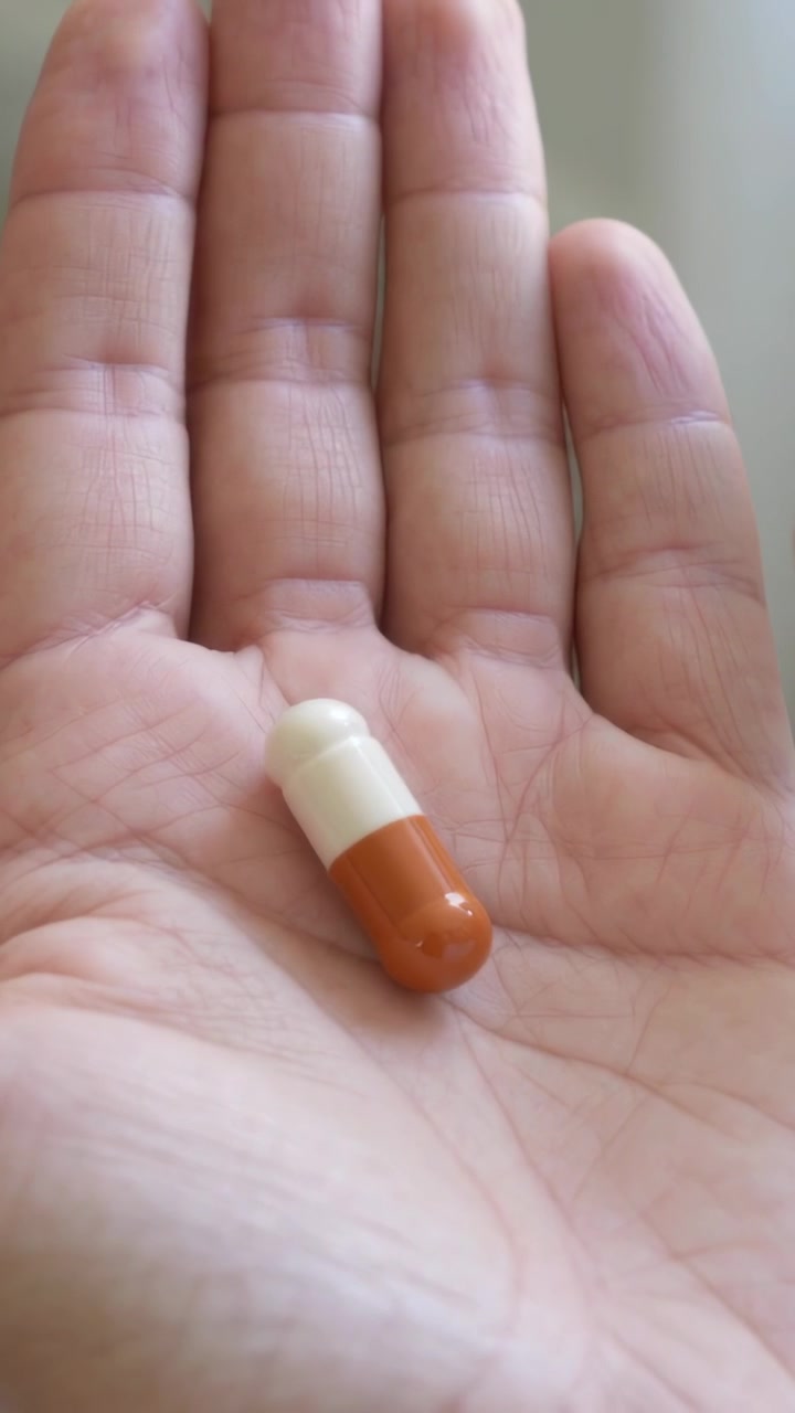 Video verticale: Mano che tiene, pollice che spinge delicatamente una capsula arancione bicolore verso il palmo, nella stanza, mentre ispeziona