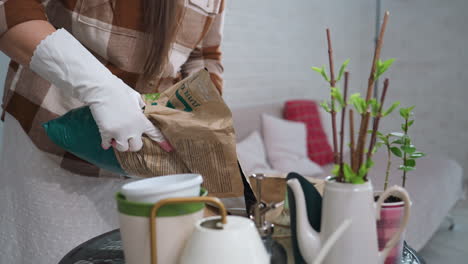 woman wearing white gloves with pink fingertips pours black sand from cut open bag into wooden container on glass table surrounded by potted winter plants against white brick wall indoors cozy
