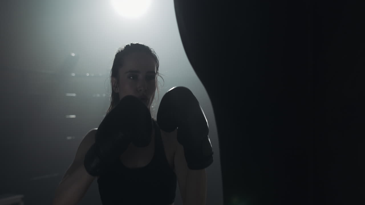 Pugilessa che si allena con un sacco da boxe in una palestra buia e fumosa.