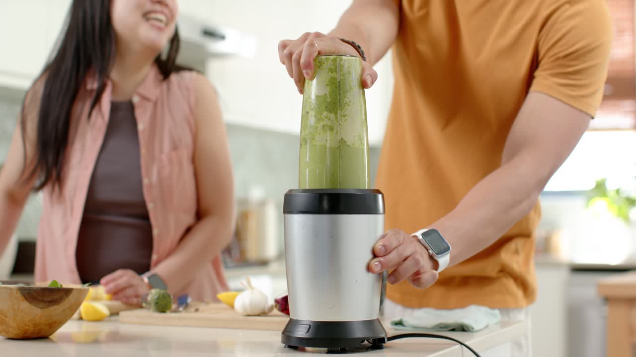 Asiatisk par lager grønn smoothie ved kjøkkenøya, mannen plasserer en kopp på basen og betjener blenderen
