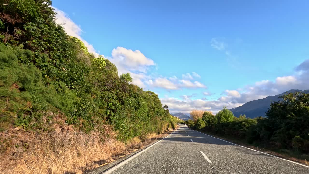 Vehicle drives along sunlit rural highway bordered by greenery, mountains, and blue sky
