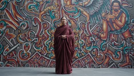 buddhist monk in maroon robes standing with hands clasped in front of a vibrant mural featuring intricate designs and ancient deities, embodying spiritual tranquility and artistic expression