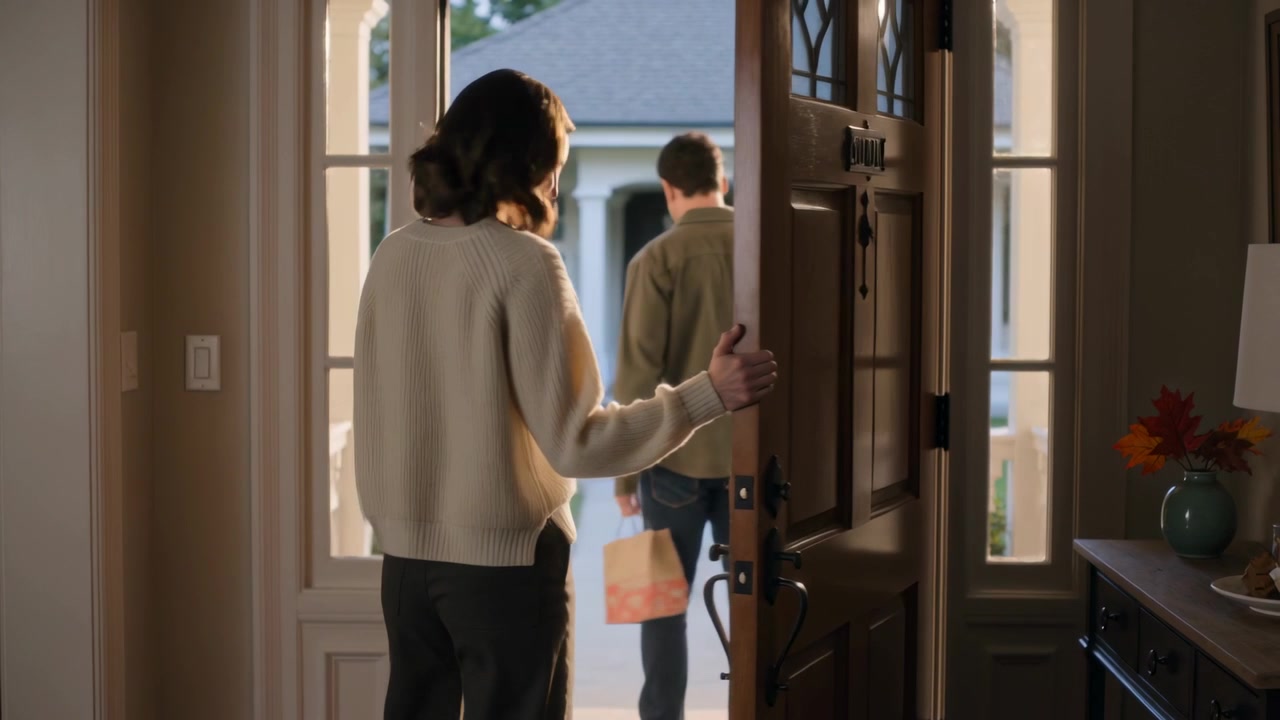 Aprendo la porta d'ingresso di casa, una donna in maglione osserva un uomo con una borsa che cammina, dando un'occhiata e chiudendo la porta.