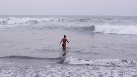 Woman-in-teeny-bikini-thong-walks-into-grey-Pacific-Ocean-waves