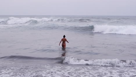 Mujer-En-Diminuto-Bikini-Tanga-Camina-Hacia-Las-Grises-Olas-Del-Océano-Pacífico