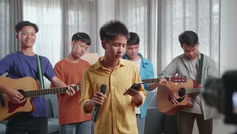 group of young asian friends live stream at home, a boy singing and reading the lyrics on smartphone while his friends playing musical instruments