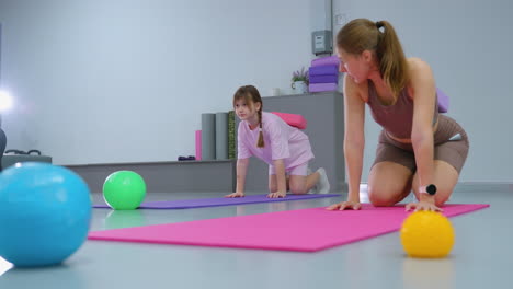 gym supervisor kneeling on mat guiding young girl performing same movement, various colorful exercise balls scattered on floor in bright fitness space with gym equipment in background