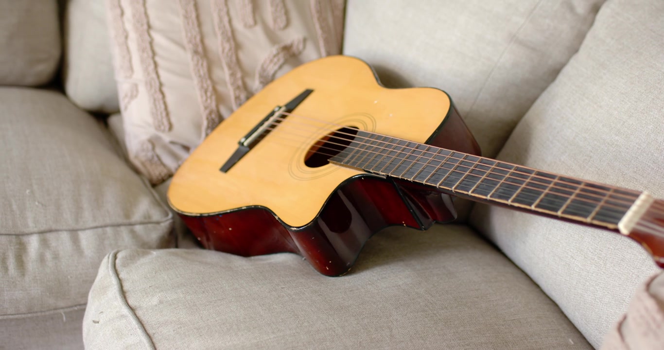 La cámara ajusta el encuadre y el enfoque, acercándose y centrando una guitarra acústica en un sofá beige
