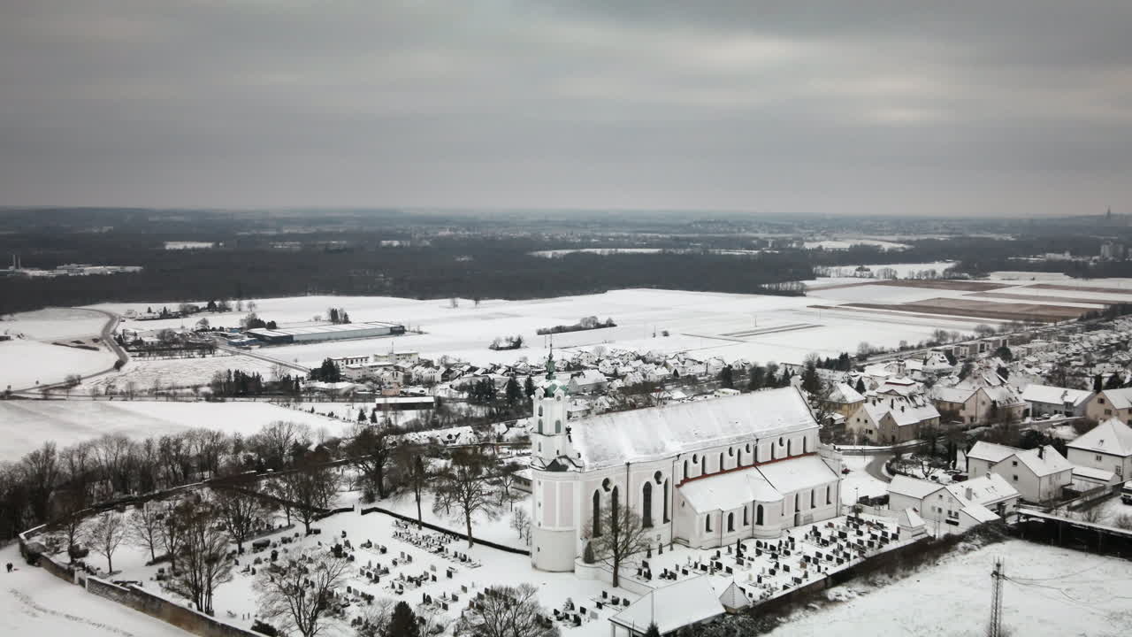 Volo in avanti di un drone verso la chiesa innevata di Elchingen tra case di paese, cimitero, campi innevati, strade, colline. Perfetto per documentari di viaggio, annunci pubblicitari, rivelazioni