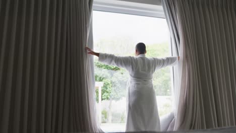 Feliz-Mujer-Afroamericana-De-Talla-Grande-Con-Bata,-Cortinas-Y-Mirando-Por-La-Ventana