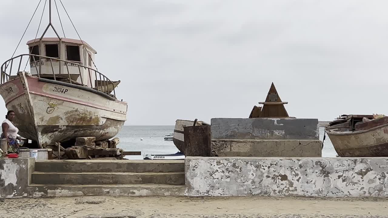 Aged fishing boat on the shore in Sal Rei, Cape Verde, with local children and a man nearby. The scene reflects the rustic coastal charm and daily life in an African island village