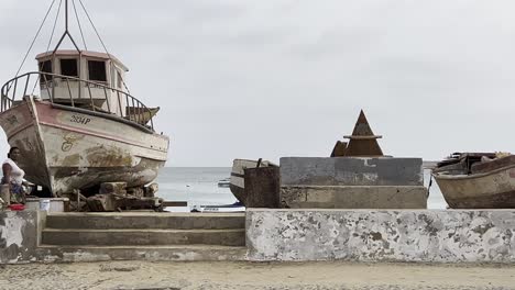 aged fishing boat on the shore in sal rei, cape verde, with local children and a man nearby. the scene reflects the rustic coastal charm and daily life in an african island village