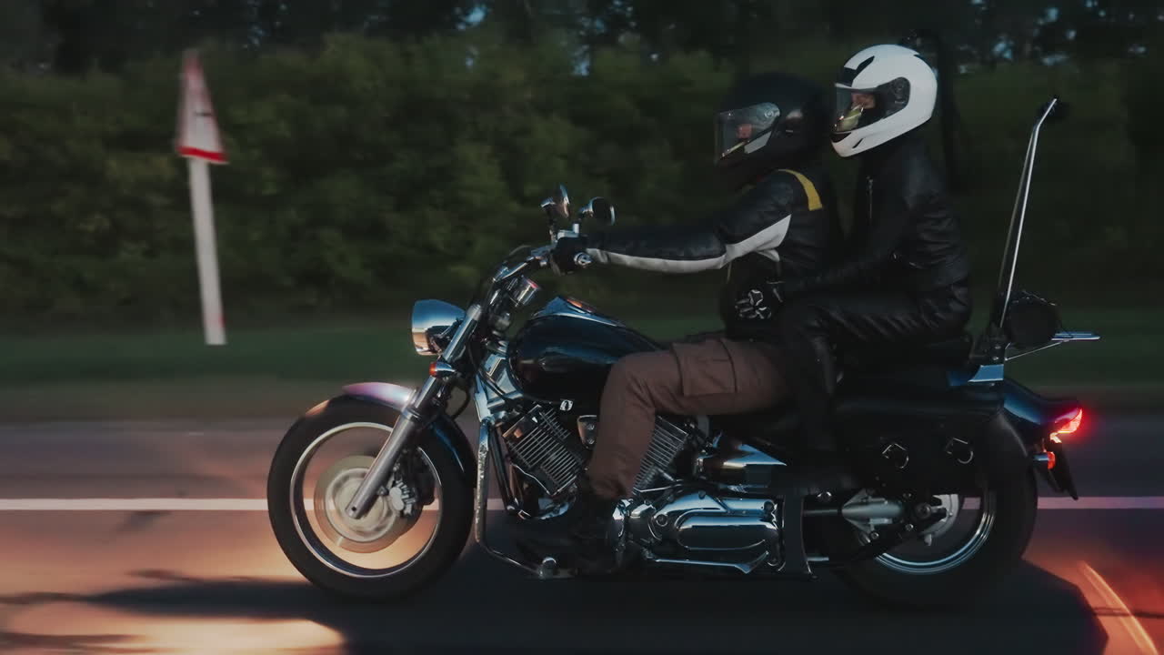 Side view of motorcyclist with passenger riding along highway during evening trip, both wearing helmets and leather jackets, motorcycle reflecting road lights while moving past green roadside trees