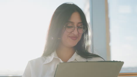 close up happy woman shaking head and smiling while operating tablet with sunlight glow illuminating hair and face against blurred window and city background