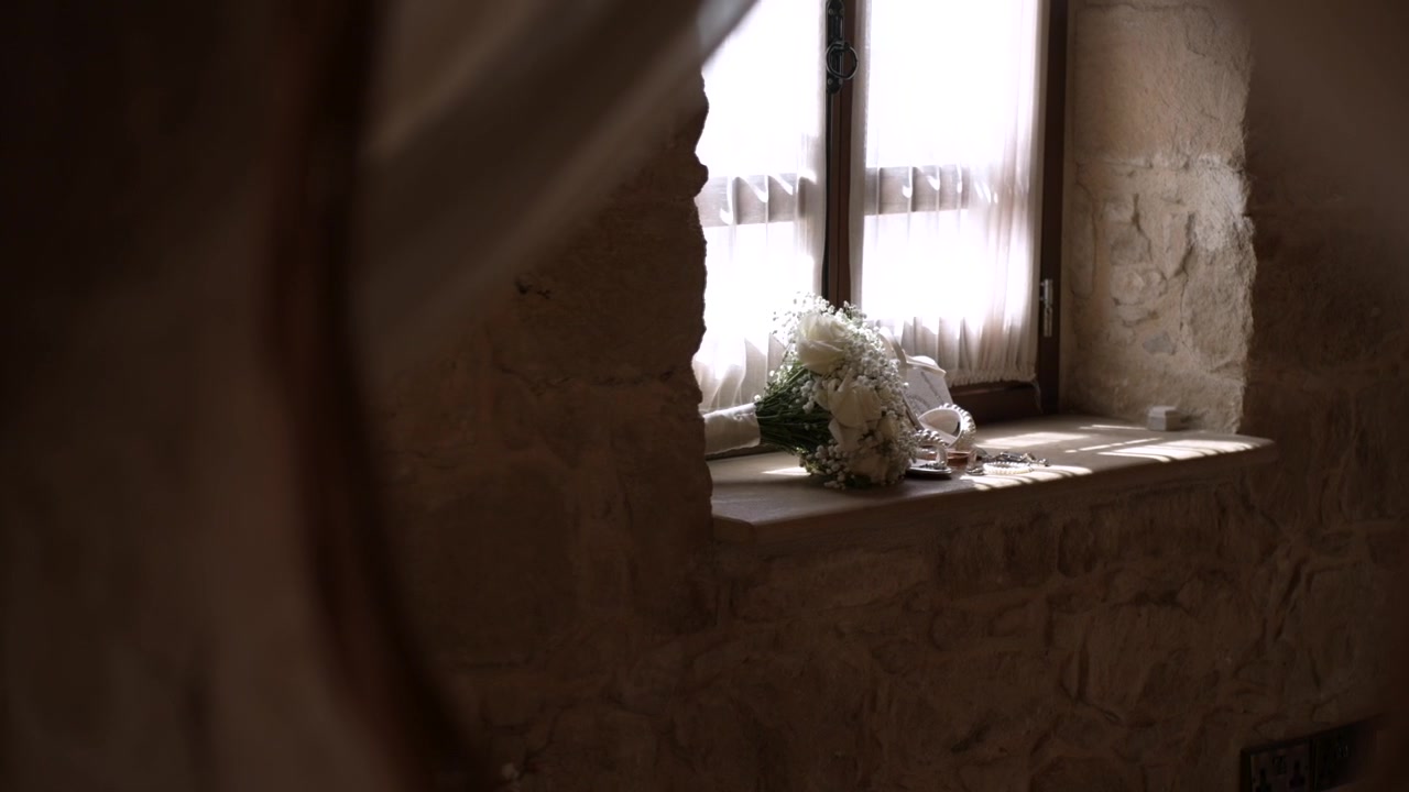 A bouquet of flowers sits on a windowsill in a rustic room. Soft natural light enters through the window. This moment captures the quiet preparation for a wedding.