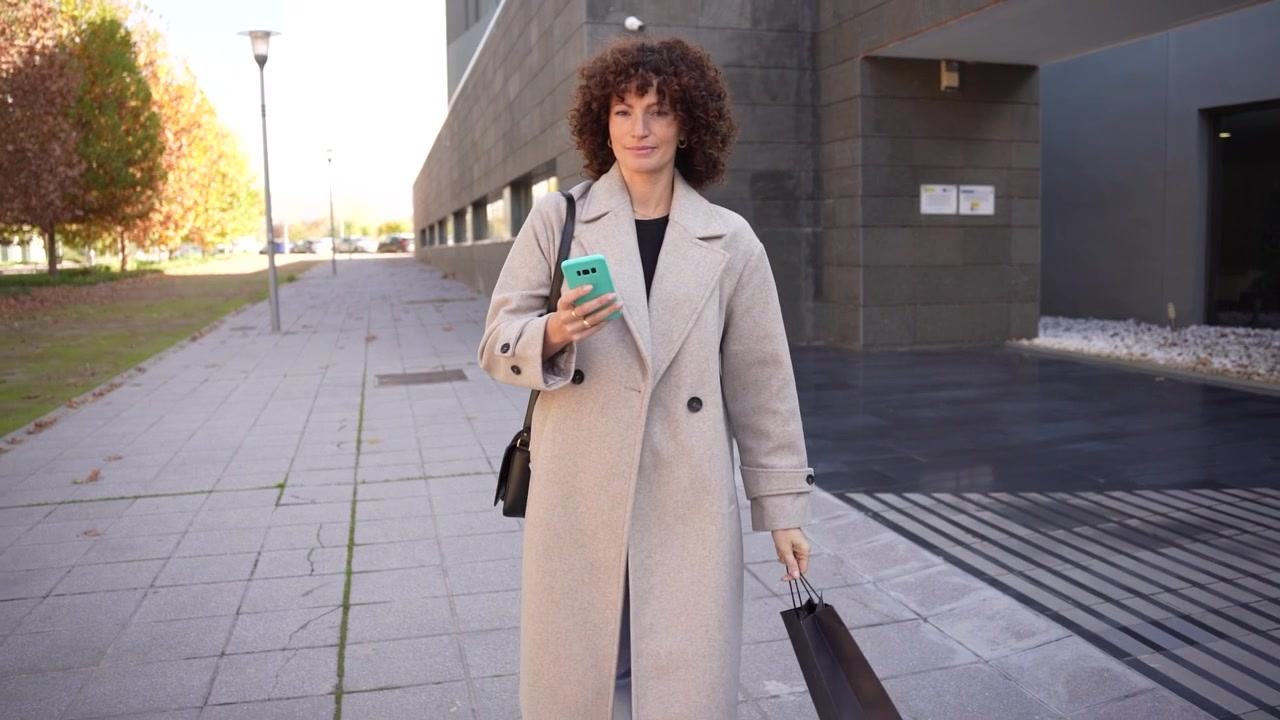 Una donna alla moda che indossa un outfit di tendenza sta camminando per una strada urbana mentre usa il suo smartphone.