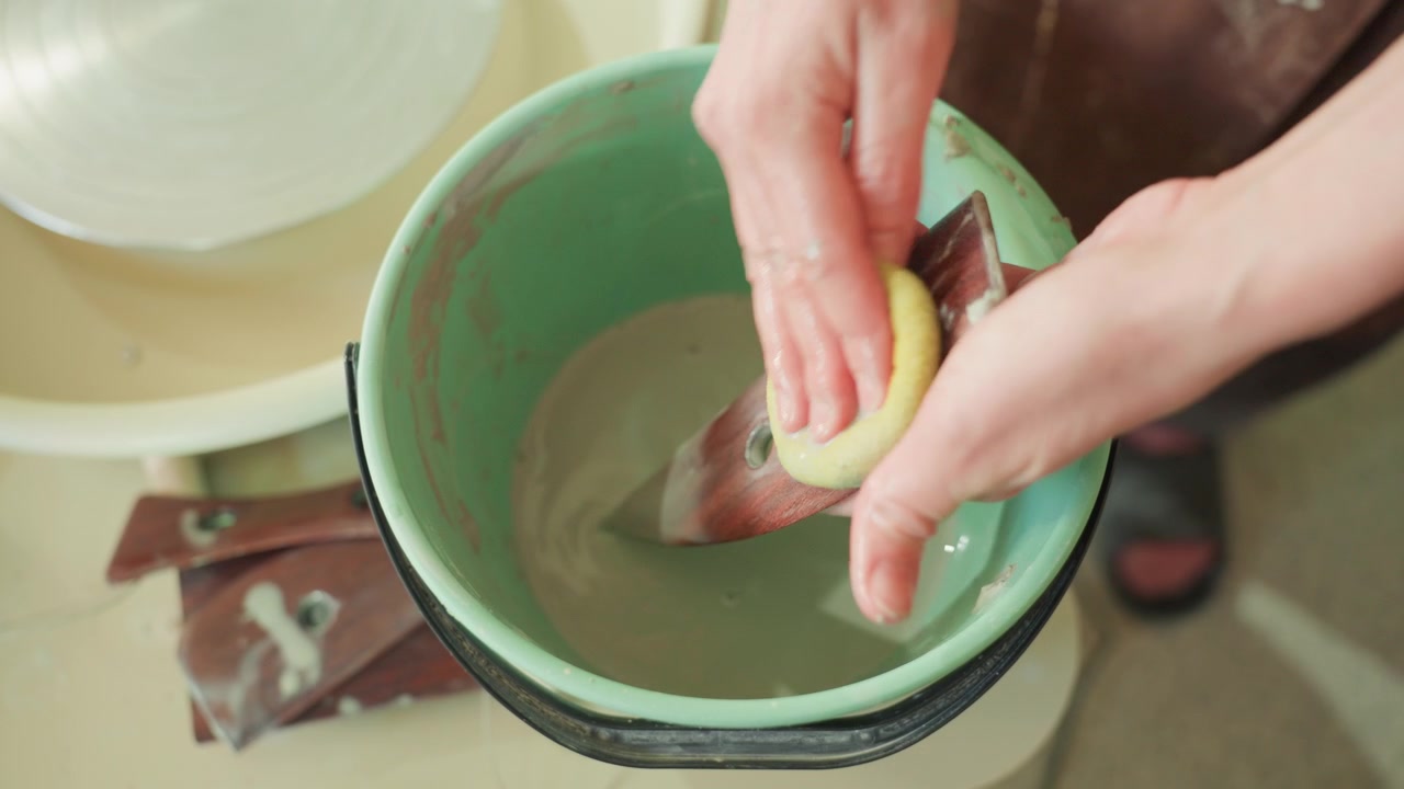 Hands cleaning pottery tool in bucket, closeup of sponge and metal rib by wheel. Quiet studio scene showing artisan rinsing clay residue, gentle circular motions, green basin and wet hands, mindful