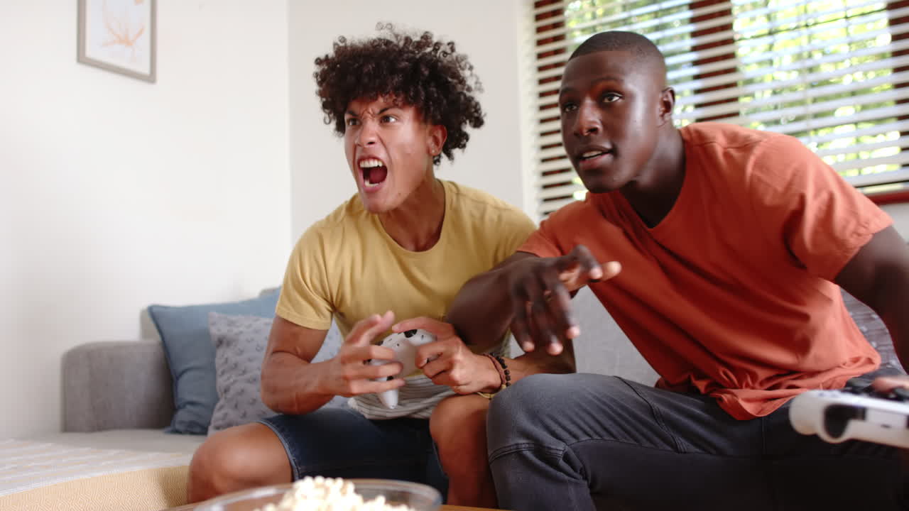 Playing video games, two multiracial male friends sitting on couch, enjoying leisure time, at home