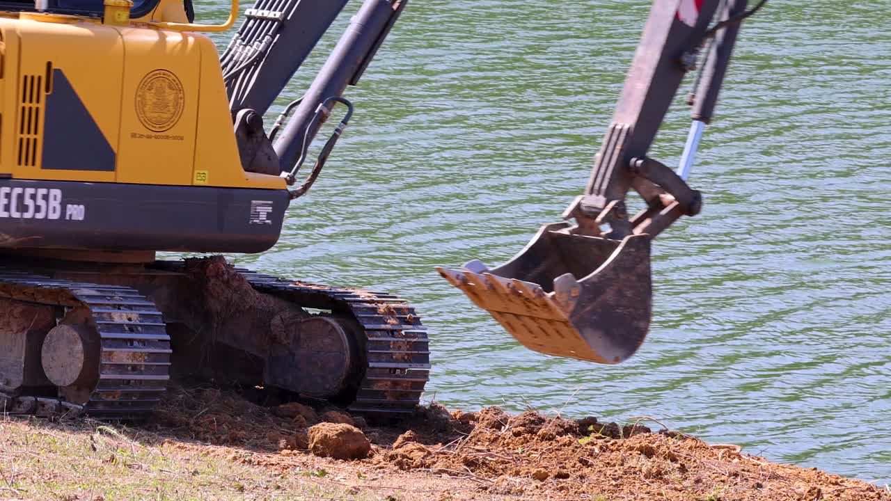 Yellow excavator digs earth by lakeside in daylight, camera steady, natural outdoor environment