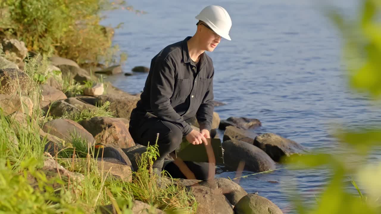 Worker in helmet crouching by riverbank testing water with clipboard in sunlight