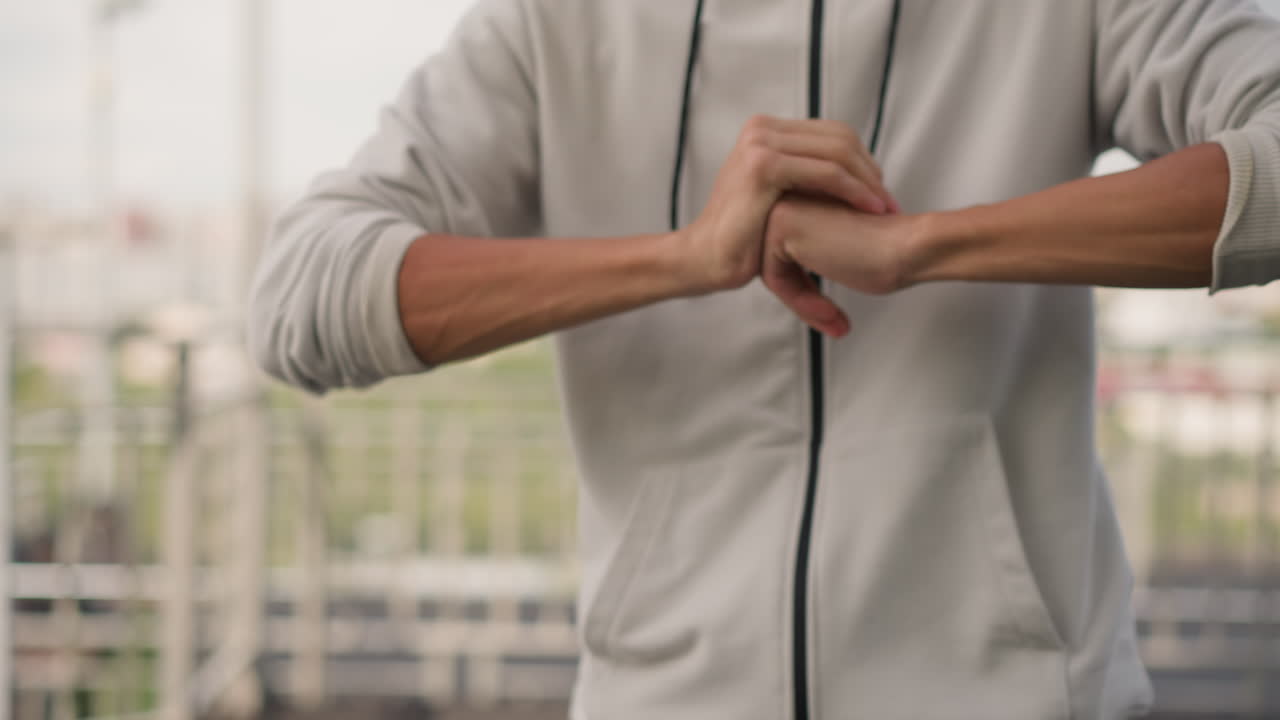 옥상 난간에서 소매를 걷어 올리는 남성, 손목과 주먹 클로즈업, 지퍼가 달린 후드티, 흐릿한 도시 스카이라인이 배경으로 보이고, 흐린 날씨 아래 훈련이나 대결을 암시하는 강렬하고 집중된 자세