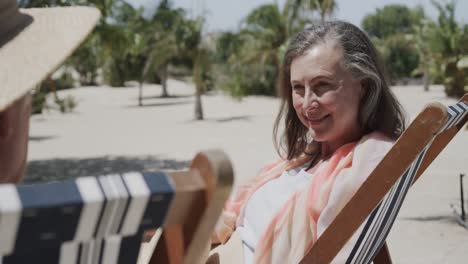 una feliz pareja caucásica sentada en sillas de sol en una playa soleada hablando en cámara lenta