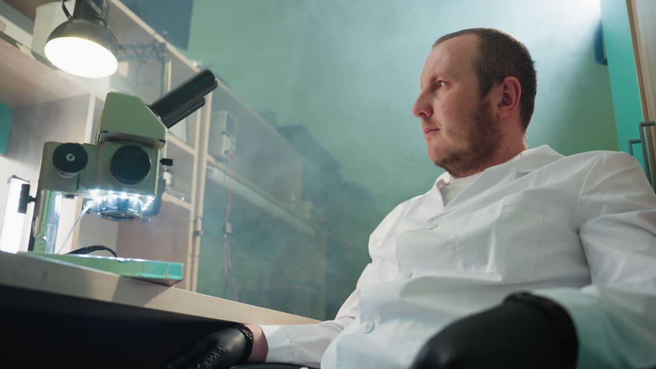 잘 장착 된 실험실에 앉아있는 실험실 코트를 입은 기술자의 근접 시각, 기술자는 초점을 맞추고 현미경으로 빛을 비추는 것처럼 보입니다.