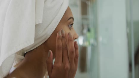 Mixed-race-woman-wearing-towel-on-head-applying-cream-on-her-face