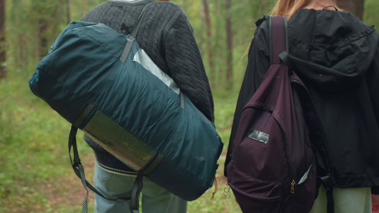 Rear view of siblings walking through forest with camping bags, one carrying large green duffel and other with purple backpack, surrounded by dense greenery and tall woodland trees