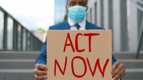 close-up view of african american elegant young man in facial mask showing act now" signboard in the street"