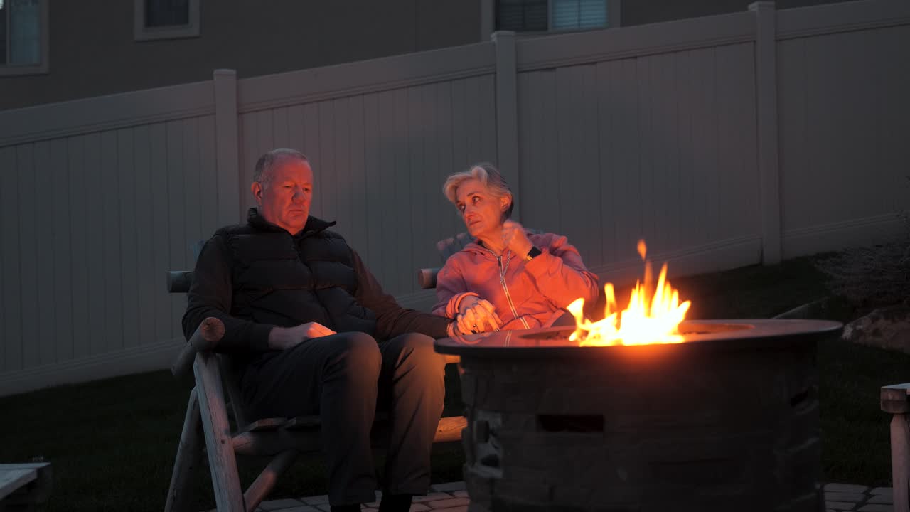 una pareja de mayores tomándose de la mano y hablando junto a un pozo de fuego en el patio trasero